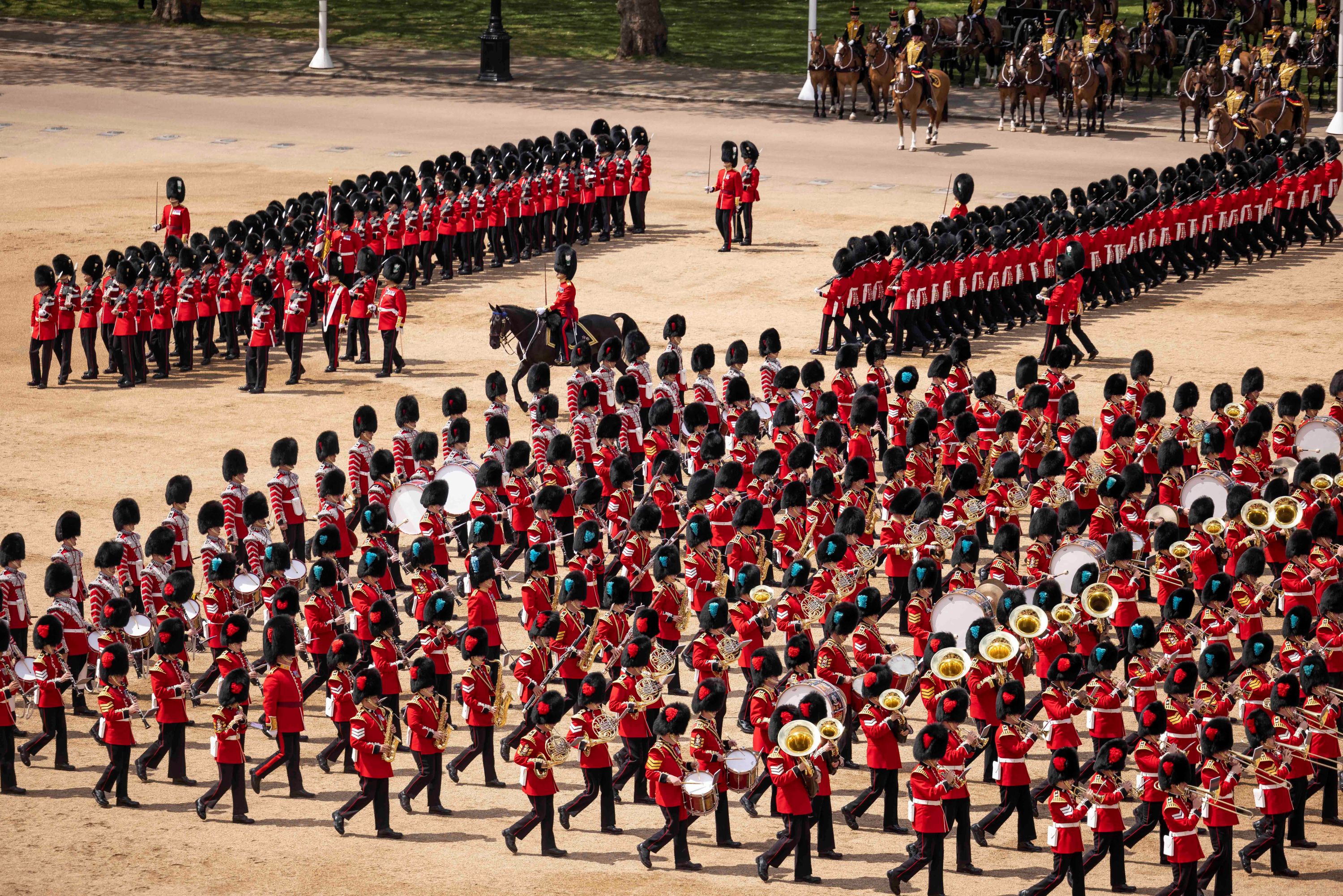 A large military parade with Guards in scarlet ceremonial uniforms. There are military musicians in the front playing brass instruments and drums. Behind them are marching infantry soldiers. In the back, there is a group of soldiers on horseback, from the King's Troop Royal Horse Artillery Regiment. 